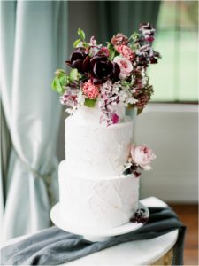 Three tier wedding cake with maroon and pink flowers on it and gold leaf